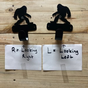Op de afbeelding: Twee zwarte metalen wandhaken in de vorm van een Bigfoot-figuur op een skateboard. De haak links is naar links gericht en de haak rechts naar rechts. De tekst op het witte papier onder de haken luidt "R = Looking Right" en "L = Looking Left".