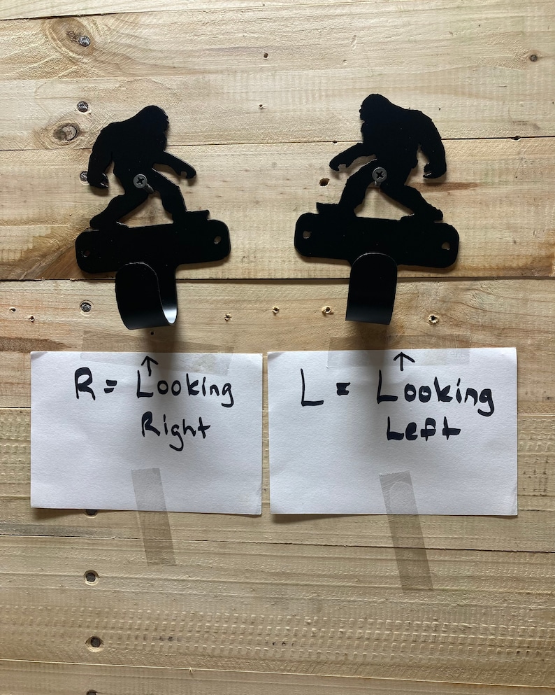 Op de afbeelding: Twee zwarte metalen wandhaken in de vorm van een Bigfoot-figuur op een skateboard. De haak links is naar links gericht en de haak rechts naar rechts. De tekst op het witte papier onder de haken luidt "R = Looking Right" en "L = Looking Left".