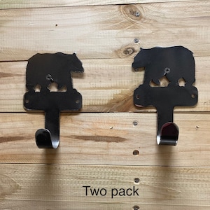 Op de afbeelding: Twee zwarte metalen haken in de vorm van een beren silhouet met een gebogen haak aan de onderkant. De haken zijn bevestigd aan een houten oppervlak. De tekst "Two pack" is zichtbaar onder de haken.