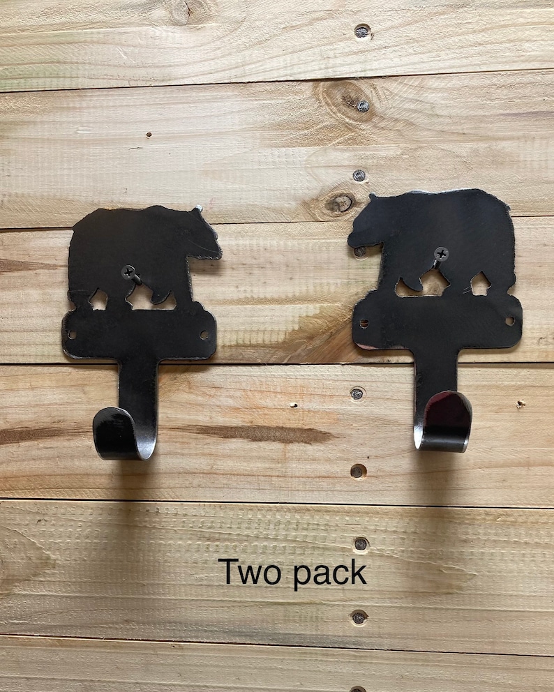 Op de afbeelding: Twee zwarte metalen haken in de vorm van een beren silhouet met een gebogen haak aan de onderkant. De haken zijn bevestigd aan een houten oppervlak. De tekst "Two pack" is zichtbaar onder de haken.