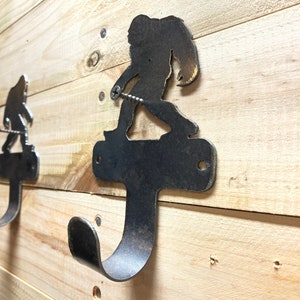 Op de afbeelding: Een metalen wandhaak in de vorm van een Bigfoot-silhouet. De haak is roestbruin en heeft een enkele schroef die hem aan een houten muur bevestigt.