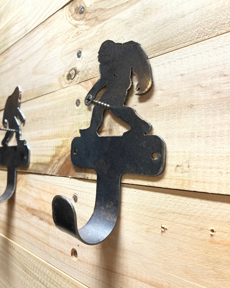 Op de afbeelding: Een metalen wandhaak in de vorm van een Bigfoot-silhouet. De haak is roestbruin en heeft een enkele schroef die hem aan een houten muur bevestigt.