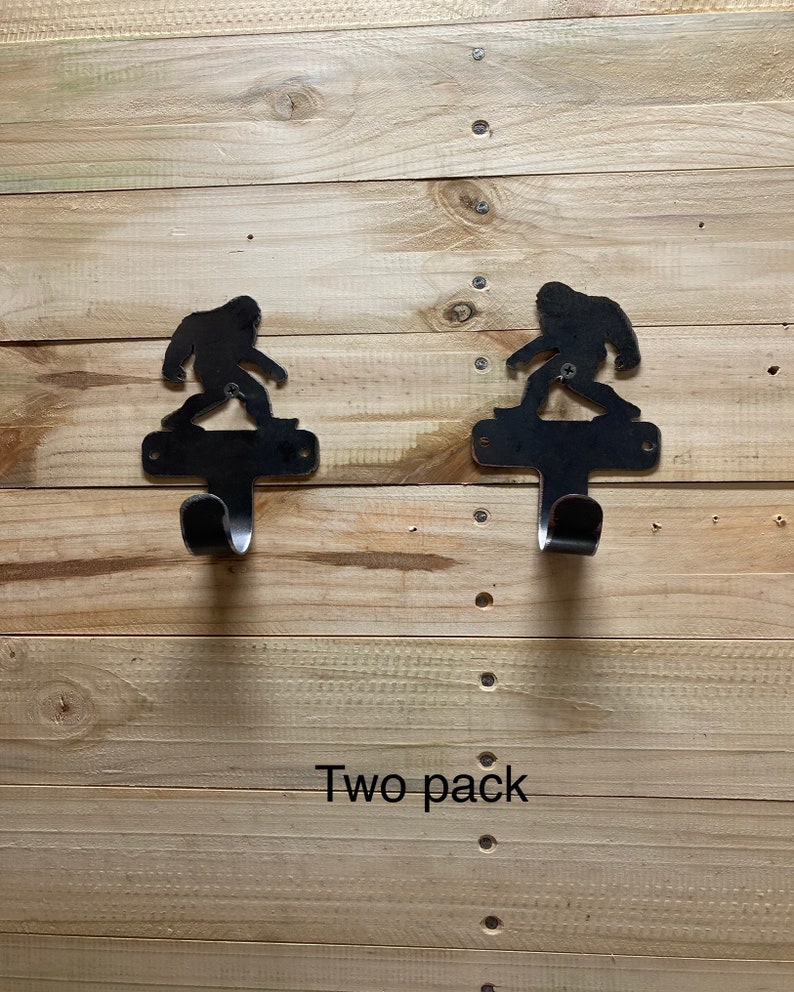Op de afbeelding: Twee zwarte metalen haken in de vorm van Bigfoot, gemonteerd op een houten muur. De haken zijn gelabeld "Two pack".