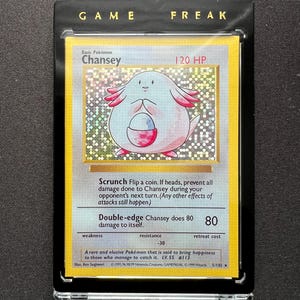 Puede incluir: Una carta coleccionable de Pokémon con Chansey, una criatura rosa con forma de huevo con brazos pequeños y una bolsa. La carta está en una funda protectora con las palabras "GAME FREAK" en la parte superior. El texto de la carta incluye "Scrunch" y "Double-edge".