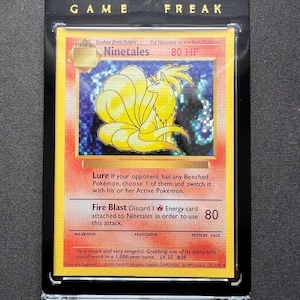 Puede incluir: Una carta Pokémon Ninetales en un estuche protector negro. La carta presenta una criatura amarilla con forma de zorro y nueve colas, sobre un fondo colorido. El texto "GAME FREAK" está en la parte superior.