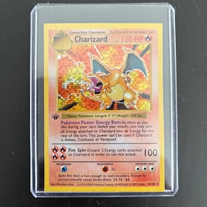 Puede incluir: Una carta coleccionable de Pokémon Charizard en una funda protectora. La carta presenta un dragón naranja llameante con las alas extendidas. El texto de la carta incluye el nombre "Charizard", los HP y los detalles de ataque. La carta mide 170 cm de largo y pesa 90 kg.