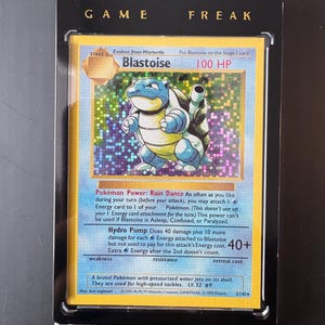 Puede incluir: Una tarjeta coleccionable de Pokémon enmarcada con Blastoise, una criatura azul con forma de tortuga. La tarjeta tiene un fondo holográfico y texto que incluye "100 HP" e "Hydro Pump". El marco tiene las palabras "GAME FREAK".