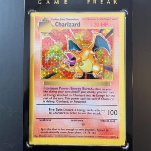 Könnte beinhalten: Eine Charizard Pokémon-Karte in einer schwarzen Schutzhülle. Die Karte zeigt eine lebendige Illustration von Charizard mit Text wie "Charizard", "120 HP" und Spielanweisungen. Auf der Hülle steht oben "GAME FREAK".