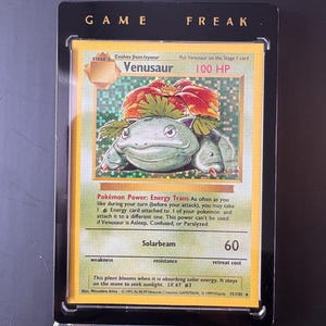May include: A Venusaur Pokémon trading card, stage 2, with 100 HP, in a black display case. The card features a green and red illustration of Venusaur. The text "GAME FREAK" is at the top.