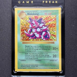 Puede incluir: Una tarjeta coleccionable de Pokémon enmarcada con Nidoking, una criatura morada y rosa. La tarjeta tiene un fondo holográfico y el texto "Nidoking 90 HP". El marco tiene las palabras "GAME FREAK" en letras doradas.