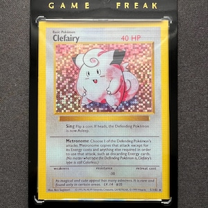 Puede incluir: Una carta coleccionable de Pokémon Clefairy en una funda protectora. La tarjeta presenta una ilustración de Clefairy rosa y blanca con texto que incluye "GAME FREAK" y "Basic Pokémon Clefairy 40 HP".