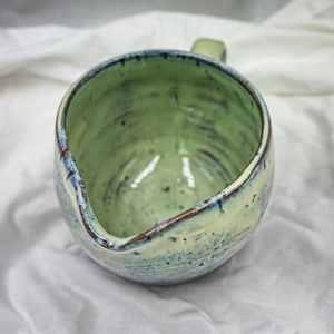 Può includere: Brocca in ceramica fatta a mano con corpo arrotondato e piccolo manico. L'interno è di colore verde chiaro, con un bordo verde scuro e blu. L'esterno ha una finitura macchiettata e strutturata.
