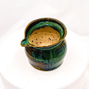 May include: A small, handmade ceramic pitcher with a spout. The pitcher features a glossy glaze in shades of green, brown, and blue. The interior is a speckled tan color. The rim and spout are highlighted with a darker green.