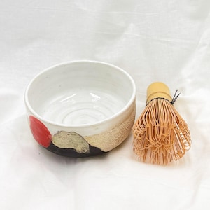 Può includere: Una ciotola per matcha in ceramica bianca con un accento rosso e una fascia beige strutturata. Accanto alla ciotola c'è un frullino per matcha in bambù. La ciotola ha una base nera ed è progettata per preparare il tè matcha.