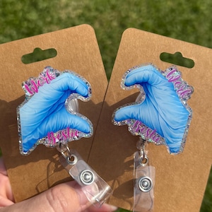 May include: Two badge reels featuring a glittery, heart-shaped design formed by two blue medical gloves. The text Work Bestie is printed in pink on each piece. The badge reels are attached to brown cardboard display cards with a top hanging hole.