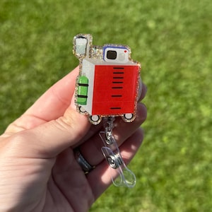 May include: A badge reel featuring a glittery acrylic topper shaped like a medical crash cart. The cart is red with a white top, a green oxygen tank on the side, and a monitor screen on top. The reel includes a clear plastic strap and metal clip.