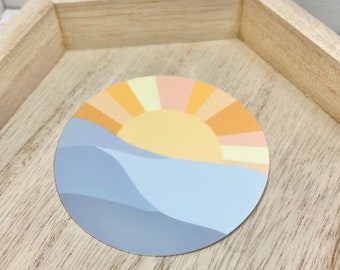 May include: A round sticker with a blue, yellow, and orange sunset design. The sticker features a stylized sun with rays and a wavy blue line representing the horizon.
