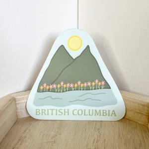 Może przedstawiać: Naklejka przedstawiająca górski krajobraz z żółtym słońcem, zielonymi górami i rzędem różowych i żółtych kwiatów. Tekst "BRITISH COLUMBIA" jest wydrukowany na dole na zielono.