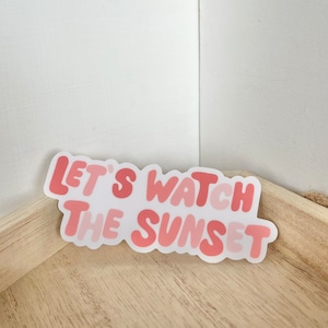 May include: Pink and white sticker with the text "Let's watch the sunset" in a retro font.