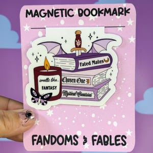 Può includere: Segnalibro magnetico a tema fantasy. Il design presenta una pila di libri viola con titoli come "Fated Mates" e "Mystical Creatures", una candela, una farfalla e una spada. È presente anche il testo "Magnetic Bookmark" e "Fandoms & Fables".