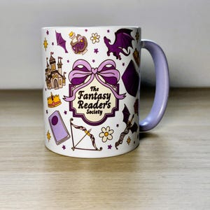 Może przedstawiać: Biały ceramiczny kubek z fioletowym uchwytem i obramowaniem. Kubek jest ozdobiony ilustracjami o tematyce fantasy, w tym zamkiem, smokiem i łukiem ze strzałą. Tekst "The Fantasy Reader's Society" jest wyraźnie widoczny na kubku.