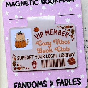 Könnte beinhalten: Ein magnetischer Lesezeichen mit hellviolettem Hintergrund und weißen Sternen. Das Lesezeichen zeigt den Text "MAGNETIC BOOKMARK" und "FANDOMS & FABLES". Es hat auch eine VIP-Mitgliedskarte mit einer Katze in einer Kürbisgrafik und dem Text "Cozy Vibes Book Club".