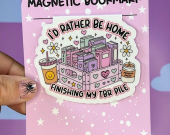 May include: A pink magnetic bookmark with the text "MAGNETIC BOOKMARK" at the top. The bookmark features a graphic of books in a crate, a drink, and a candle. The text "I'D RATHER BE HOME FINISHING MY TBR PILE" is also included. The bottom of the bookmark reads "FANDOMS & FABLES".