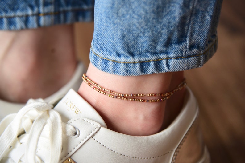 May include: A close-up of a person's ankle wearing a delicate gold anklet with small, multi-colored beads. The anklet is layered, creating a subtle and stylish look.