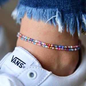 May include: A double-strand anklet made of small, multicolored glass seed beads in shades of pink, blue, orange, white, and gold. The anklet is worn around the ankle above a white sneaker and below the frayed hem of blue denim jeans.