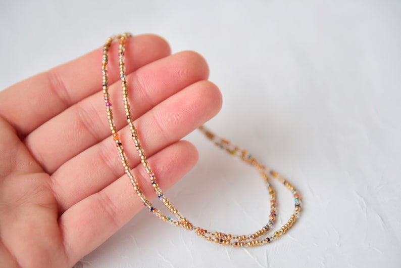 May include: A delicate beaded necklace featuring small metallic gold beads interspersed with tiny, multicolored glass seed beads. The strand is thin and flexible, showcasing a repeating pattern of gold and vibrant accent colors throughout.