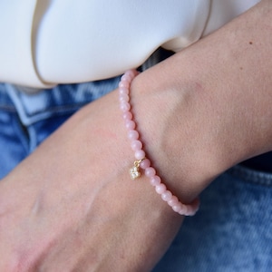 May include: A delicate pink beaded bracelet with a small gold charm featuring a clear stone. The bracelet is worn on a wrist, with a white shirt and blue jeans in the background. The bracelet is a simple and elegant piece of jewelry.