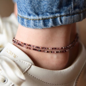 May include: Two beaded anklets in shades of burgundy, pink, and brown encircle an ankle. The anklets are worn over a white sneaker and under blue denim jeans. The beads appear to be small and delicate, creating a subtle, elegant accessory.