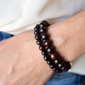 May include: Two dark brown beaded bracelets are worn on a wrist. The bracelets are made of round, polished beads. The skin tone is light, and the person is wearing a white shirt and blue jeans.