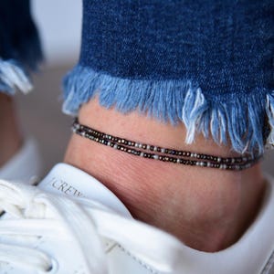 May include: Close-up of an ankle adorned with a delicate beaded anklet. The anklet features small, dark beads and silver-toned beads. The ankle is positioned above a white sneaker and below frayed denim jeans.