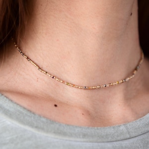May include: Delicate beaded necklace with a gold chain and small, colorful beads. The necklace is worn around the neck, resting on a person's skin, and is paired with a gray shirt. The necklace is a choker style.