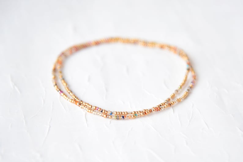 May include: Delicate beaded necklace with a gold-tone chain and colorful seed beads. The necklace is composed of two strands, creating a layered effect. The beads feature a mix of colors, including gold, orange, and purple, on a white textured background.