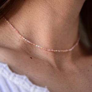 May include: A delicate choker necklace made with small pink and white beads. The necklace is worn by a person with a white shirt.
