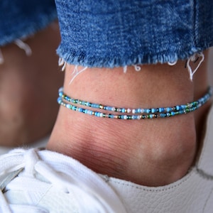 May include: Two beaded anklets in shades of blue, green, and gold are worn on an ankle. The anklets are made of small beads strung together. The person is wearing blue jeans and white sneakers.
