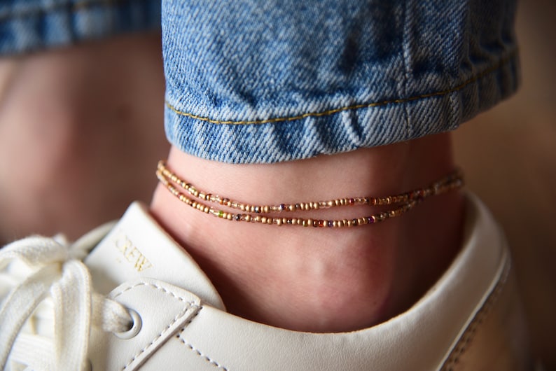 May include: A double-strand anklet made of small, metallic gold-toned seed beads with occasional multicolored accents. The anklet is worn around the ankle, positioned above a white leather sneaker and below blue denim jeans.