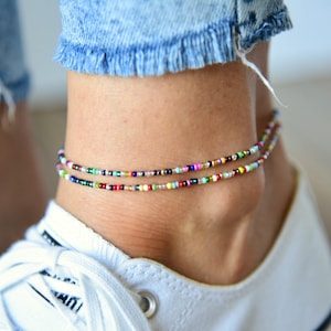 May include: Two colorful beaded anklets on a person's ankle. The anklets are made of small, multi-colored beads and are worn on top of a white sneaker.