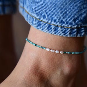 May include: Ankle bracelet featuring a delicate design with turquoise and dark green beads, interspersed with a few white pearls. The bracelet is worn on an ankle, with blue denim jeans visible in the background.