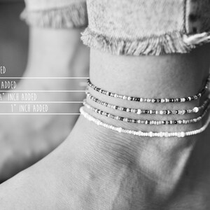 May include: Black and white image of an ankle adorned with four beaded anklets. The anklets vary in design, with beads in white, black, and silver. Text indicates measurements: 1/4 inch, 1/2 inch, 3/4 inch, and 1 inch added.
