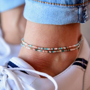 May include: A close-up of a person's ankle wearing two delicate beaded anklets. The anklets are made of small, colorful beads in shades of turquoise, pink, white, and gold. The anklets are layered on top of each other, creating a stylish and bohemian look.