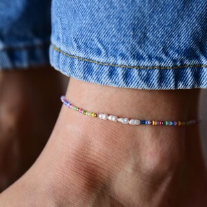 May include: Ankle bracelet featuring a delicate design with small, colorful beads and several white pearls. The bracelet is worn on an ankle, with blue denim jeans visible in the background.