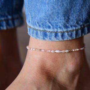 May include: Delicate beaded anklet featuring a mix of small, light pink, and white beads, interspersed with tiny gold-colored beads. The anklet is worn on an ankle, with blue denim jeans visible in the background.