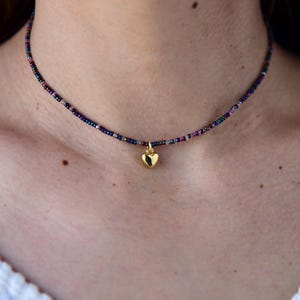 May include: A beaded choker necklace with a gold heart pendant. The necklace features small, colorful beads in shades of purple, blue, and pink. The gold heart pendant hangs delicately in the center.