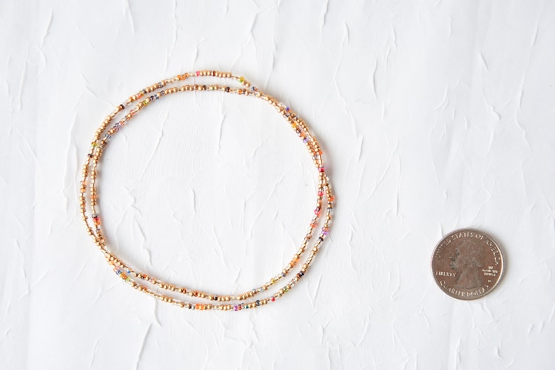 May include: A delicate beaded necklace or bracelet composed of small metallic gold beads interspersed with tiny multicolored glass beads, arranged in a circular shape next to a United States quarter coin for scale.