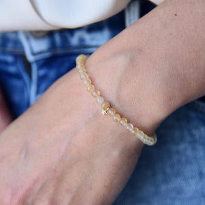 May include: A delicate bracelet featuring small, light yellow beads strung together. A small gold-colored charm with a star design is centered on the bracelet. The bracelet is worn on a wrist, with blue denim jeans in the background.