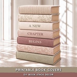 May include: A stack of seven books with textured covers in shades of beige, cream, mauve, and sage green. Three books feature printed text reading A NEW CHAPTER BEGINS, while others display subtle leaf patterns or solid fabric-like textures.