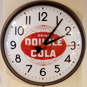 May include: A round, black-framed clock with white face and black hands. The clock face features a red oval with the text "Drink Double Cola" and a globe design.